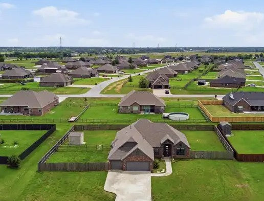 Aerial view of a suburban neighborhood with single-family homes, green lawns, paved streets, and a blue sky with clouds. 